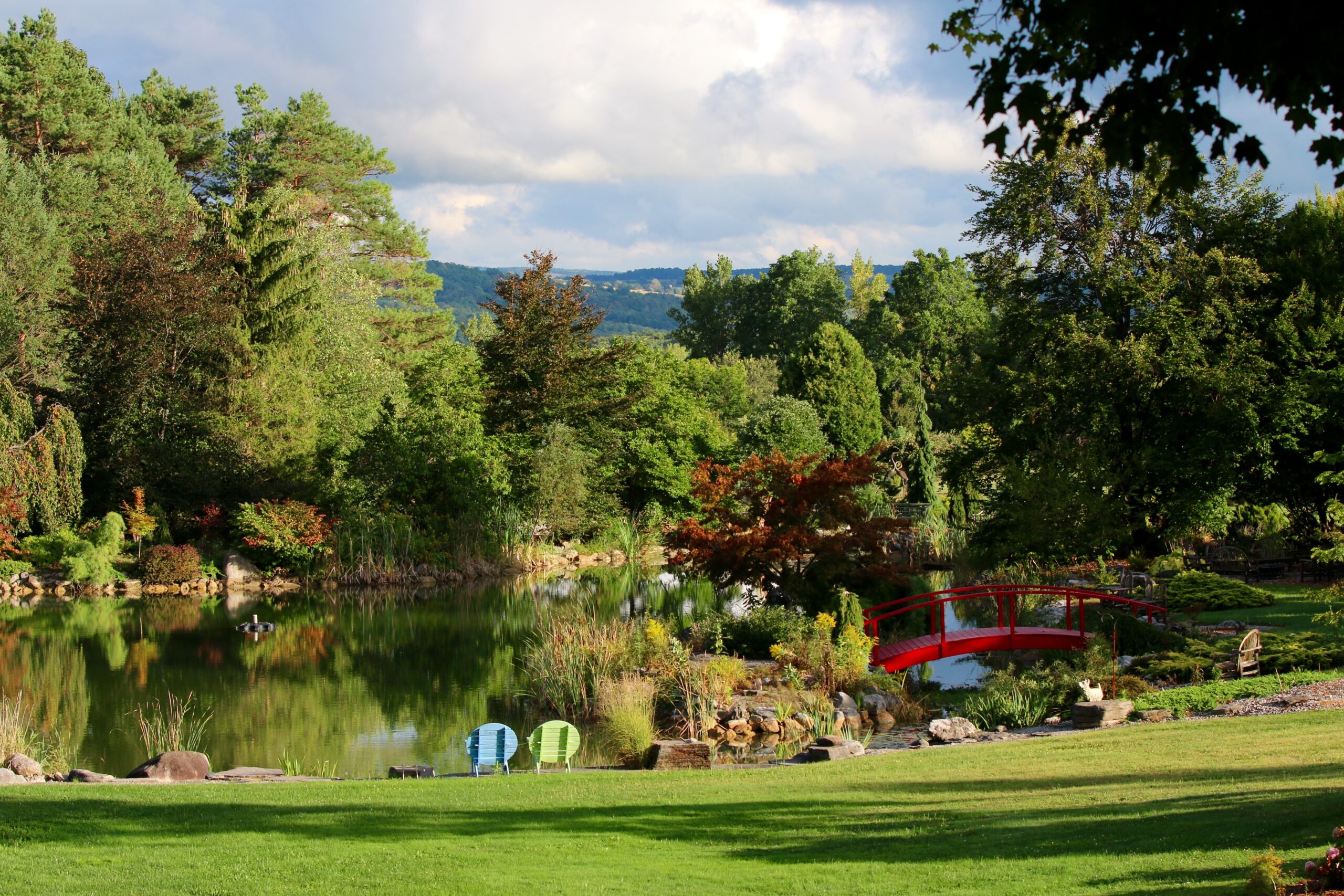 Green lawn, blue sky, a myriad of trees, and a pond with Adirondack chairs.